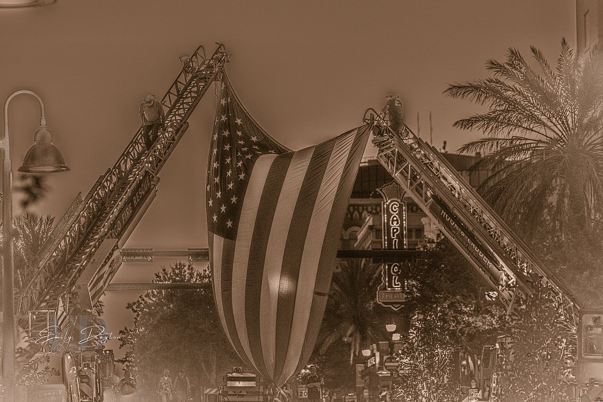 Sepia-toned photo of two ladder fire trucks with an American flag hanging between them. A man walks down the left ladder, while another man stands at the top of the right ladder. The Capitol Theatre is visible in the background.